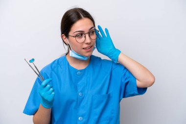 Beyaz arka planda izole edilmiş aletleri tutan beyaz dişçi kadın bir şey dinlerken elini kulağına koyar.