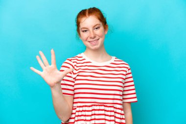 Mavi arka planda izole edilmiş genç kızılımsı kadın parmaklarıyla beşi sayıyor