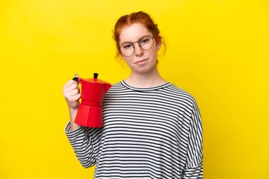 Kırmızımsı bir kadın elinde kahve demliği tutarken üzgün bir ifadeyle sarı arka planda izole edilmiş.