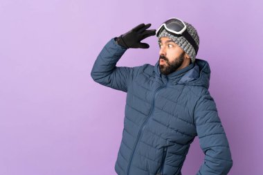 Snowboard gözlüklü kayakçı mor arka planda sürpriz bir ifadeyle yan tarafa bakarken