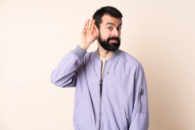 Sakallı, ceketli, soyutlanmış arka planda bir şey dinleyen, elini kulağına koyan beyaz bir adam.