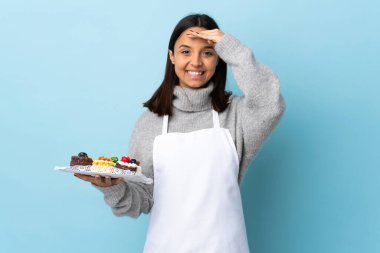 Pasta şefi, izole edilmiş mavi arkaplanda elinde büyük bir pasta tutarken mutlu bir ifadeyle selam veriyor..