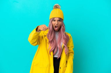 Young woman with pink hair wearing a rainproof coat isolated on blue background frustrated and pointing to the front clipart