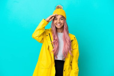 Pembe saçlı, yağmura dayanıklı bir ceket giyen genç bir kadın mavi arka planda izole edilmiş mutlu bir ifadeyle selam veriyor.