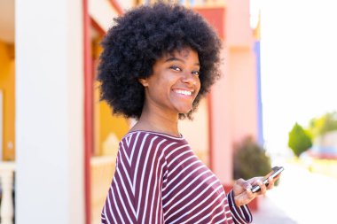 Afro-Amerikalı kız açık havada cep telefonu kullanıyor. Çok gülümsüyor.
