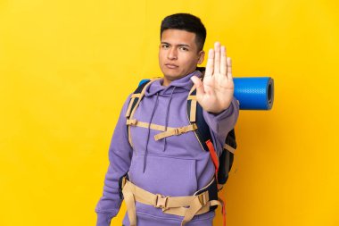 Sarı arka planda izole edilmiş büyük sırt çantalı genç dağcı dur hareketi yapıyor.