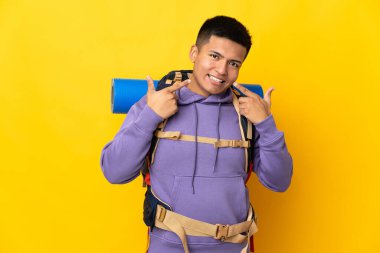 Sarı arka planda izole edilmiş büyük bir sırt çantası olan dağcı genç adam baş parmak işareti yapıyor.