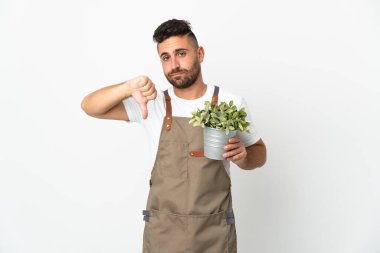 Bahçıvan, beyaz arka planda bir bitkiyi tutarken olumsuz bir ifadeyle baş parmağını aşağı gösteriyor.