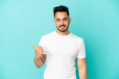 Mavi arka planda izole edilmiş genç beyaz adam bir ürünü sunmak için kenarı işaret ediyor.