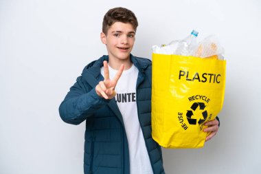 Beyaz arka planda geri dönüşüm yapmak için elinde plastik şişelerle dolu bir çanta tutan Rus ergen gülümseyerek zafer işareti gösteriyor.