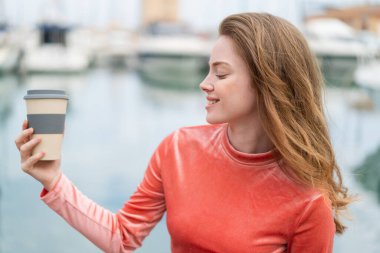 Genç kızıl saçlı kadın açık havada elinde kahveyle neşeli bir ifade ile bekliyor.