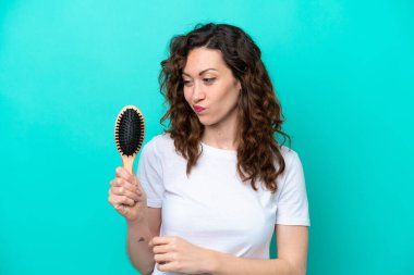 Mavi arka planda saç fırçası tutan beyaz bir kadın hüzünlü bir ifadeyle izole edilmiş.