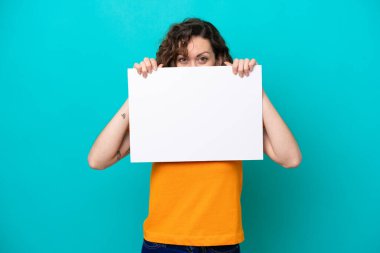 Young caucasian woman isolated on blue background holding an empty placard and hiding behind it