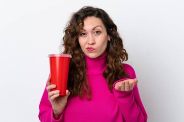 Beyaz arka planda gazoz içen genç beyaz bir kadın omuzlarını kaldırırken şüpheyle hareket ediyor.