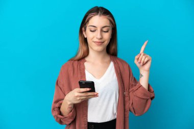 Genç beyaz kadın mavi arka planda cep telefonu ve parmak kaldırarak izole edilmiş.