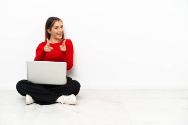 Young caucasian woman with a laptop sitting on the floor pointing to the front and smiling