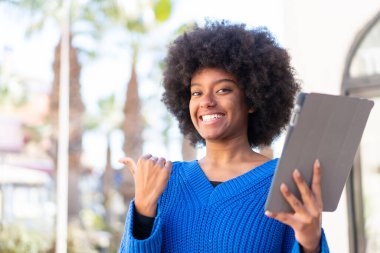 Afro-Amerikalı bir kız açık havada bir tablet tutuyor. Ürünü sunmak için yan tarafı işaret ediyor.