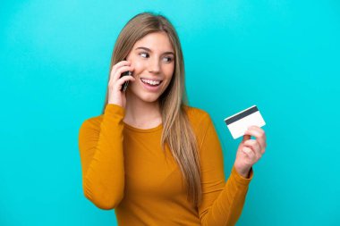 Kafkasyalı genç bir kadın mavi arka planda izole edilmiş cep telefonuyla sohbet ediyor ve elinde bir kredi kartı tutuyor.