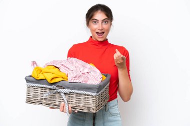 Beyaz arka planda izole edilmiş çamaşır sepeti tutan beyaz bir kadın şaşırmış ve ön tarafı işaret ediyor.