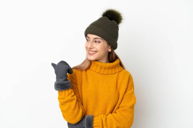 Beyaz arka planda izole edilmiş, kış şapkalı genç bir kız bir ürün sunmak için kenarı işaret ediyor.