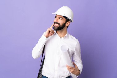 Miğferli genç bir mimar, elinde mor arkaplan üzerine planlar tutarken bir yandan da bir fikir üretiyor.