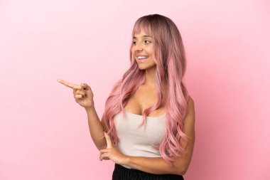 Pembe arka planda izole edilmiş pembe saçlı, melez genç bir kadın parmak göstererek bir ürün sunuyor.