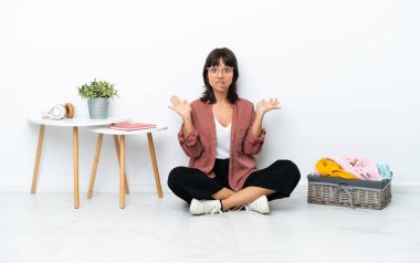 Genç melez kadın, katlanan giysiler katlıyor beyaz arka planda izole bir şekilde oturuyor ve yüzünde şok ifadesi var.