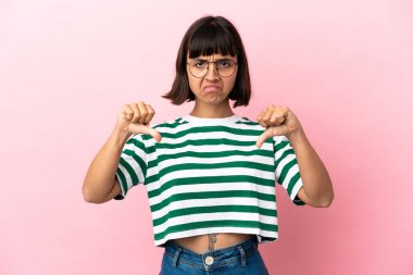 Pembe arka planda izole edilmiş genç bir ırk kadını baş parmağını iki eliyle aşağı gösteriyor.