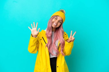 Pembe saçlı, yağmura dayanıklı bir ceket giyen genç bir kadın mavi arka planda izole edilmiş yediyi parmaklarıyla sayıyor.