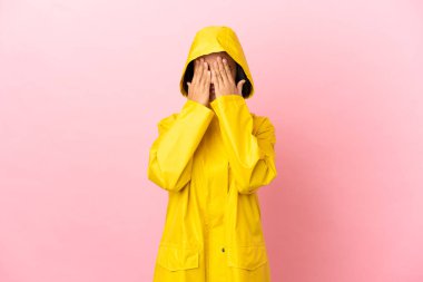 Yağmura dayanıklı bir ceket giyen genç Latin bir kadın izole arka planda gözlerini elleriyle kapatıyor.