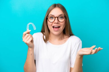 Görünmez diş telleri tutan Litvanyalı genç bir kadın mavi arka planda şok olmuş bir yüz ifadesiyle izole edilmiş.