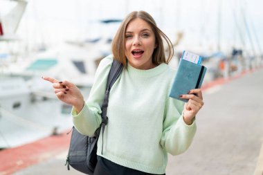 Açık havada elinde pasaport tutan genç, güzel sarışın kadın şaşırmış ve parmağıyla kenarı işaret ediyor.