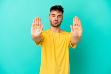 Mavi arka planda izole edilmiş genç beyaz adam dur hareketi yapıyor ve hayal kırıklığına uğruyor.