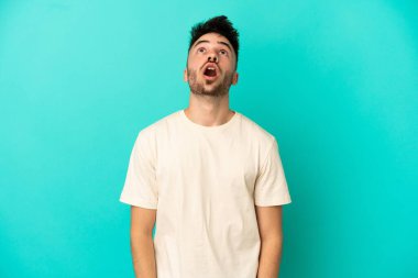 Kafkasyalı genç adam mavi arka planda izole edilmiş yukarı bakıyor ve şaşırmış bir ifadeyle