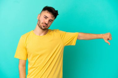 Mavi arka planda izole edilmiş genç beyaz adam olumsuz ifadeyle baş parmağını aşağı gösteriyor.