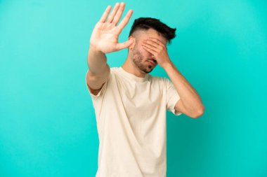 Mavi arka planda izole edilmiş genç beyaz adam jesti durduruyor ve yüzünü kapatıyor.