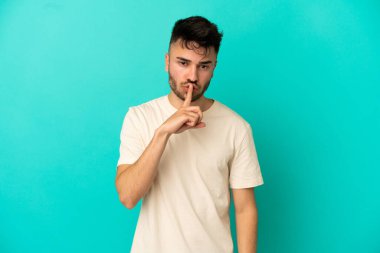 Mavi arka planda izole edilmiş genç beyaz adam sessiz bir jest işareti gösteriyor. Parmağını ağzına sokuyor.