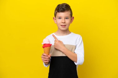 Sarı arka planda izole edilmiş kornet dondurması olan beyaz bir çocuk bir ürün sunmak için yan tarafı işaret ediyor.