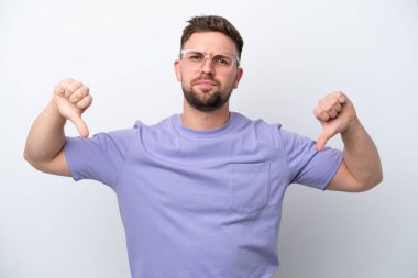 Beyaz arka planda izole edilmiş genç beyaz bir adam baş parmağını iki eliyle aşağı gösteriyor.