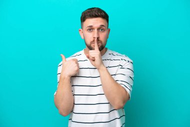 Kafkasyalı genç adam mavi arka planda izole edilmiş. Yan tarafı işaret ediyor ve sessizlik hareketi yapıyor.
