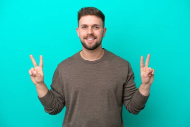 Mavi arka planda izole edilmiş genç beyaz adam iki eliyle zafer işareti gösteriyor