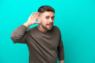 Kafkasyalı genç adam mavi arka planda izole edilmiş bir şekilde elini kulağına koyarak bir şeyler dinliyor.
