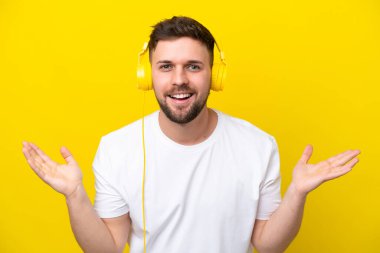 Sarı arka planda izole edilmiş genç beyaz adam şaşırdı ve müzik dinliyor.