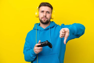 Sarı arka planda izole edilmiş bir bilgisayar oyunu kontrolörüyle oynayan genç beyaz adam, negatif ifade ile baş parmağını aşağı gösteriyor.