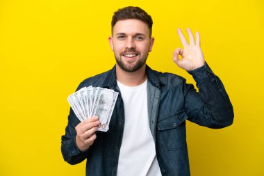 Kafkasyalı genç adam çok para alıyor. Sarı arka planda izole edilmiş. Parmaklarıyla işaret veriyor.