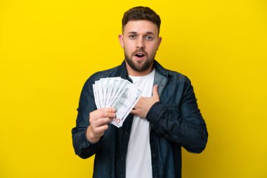 Young caucasian man taking a lot of money  isolated on yellow background surprised and shocked while looking right