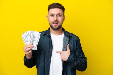 Kafkasyalı genç bir adam, sarı arka planda, şaşırtıcı bir yüz ifadesiyle izole edilmiş bir sürü para alıyor.