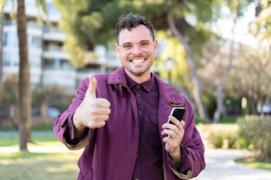 Açık havada cep telefonu kullanan genç beyaz bir adam baş parmağını kaldırıyor.