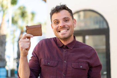 Genç, beyaz bir adam dışarıda bir cüzdan tutuyor. Şaşırmış ve şok olmuş bir yüz ifadesi var.