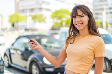 Açık havada elinde mutlu bir ifadeyle arabanın anahtarlarını tutan genç bir kadın.